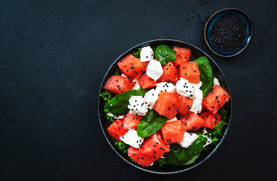 Summer Watermelon juicy salad bowl with feta cheese, spinach and black sesame seed, dark table background, top view, negative space