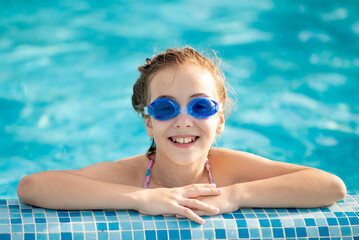 A beautiful young girl in a swimsuit bathes in a pool with blue clear water in blue swimming glasses. Summer. Rest. Vacation.