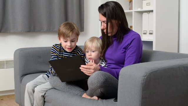 Mom with children look at the laptop monitor at home in the living room, spending time with children