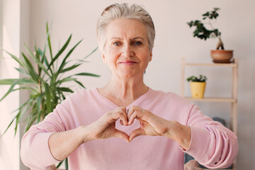 Middle aged elderlywoman sit in living room connected fingers showing heart symbol