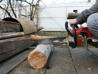 a man with an orange chainsaw in a village yard saws off a part of a dry log, a woodworker in a rural yard with a gasoline saw works with material, a peasant cuts a log in the street - obrazy, fototapety, plakaty