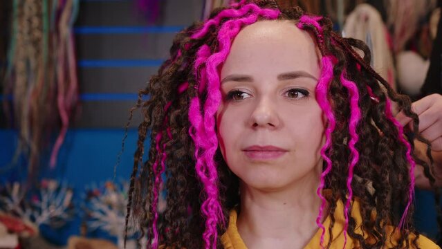 Unrecognizable person makes curly hairstyle for young woman in salon. Professional hairstylist makes pink and brown dreadlocks for client.