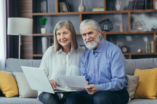 Senior Gray-haired Married Couple Man And Woman Sitting Together On Sofa At Home, Looking At Camera And Smiling, Paper Work With Laptop