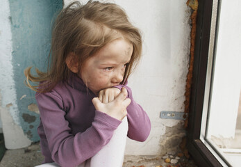 a little Ukrainian girl with a face blackened by fire, crying sitting on the floor near the window. Peaceful children and the war in Ukraine.
