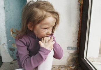 a little Ukrainian girl with a face blackened by fire, crying sitting on the floor near the window....