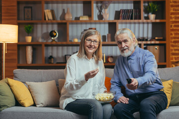 grey-haired older couple at home choose a television show to watch and switch channels with a TV...