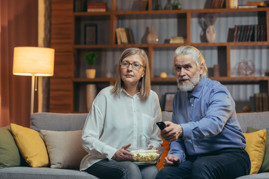 Grey-haired Older Couple At Home Choose A Television Show To Watch And Switch Channels With A TV Remote Control. Elderly Mature Family Husband And Wife Cannot Choose A Show. Watching News Or Film