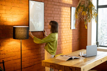 Young woman hangs and fixes photo frame on brick wall, decorating living space at home. Mockup...