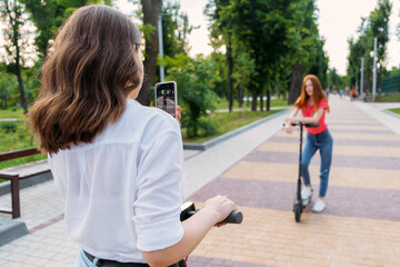 Two girl friends riding push e scooter and do selfie via their smartphones outdoors. Group gen z young people using electric scooter in city park