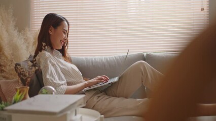 Smiling Asian freelance business women casual wear sitting on sofa using laptop looking at screen typing message,working call video conference in living room.Happy young girl relax at home in summer - Powered by Adobe
