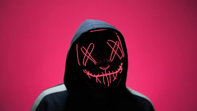 Man in creepy mask wearing hood. Mystery man with flashing light. Halloween concept at pink background. Black mask with pink led neon. 4K, UHD
