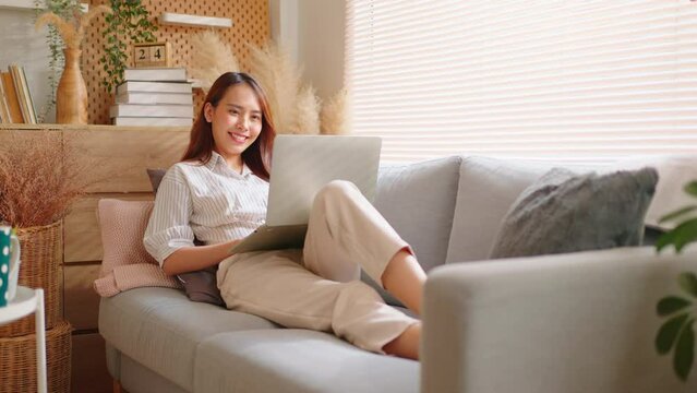 Smiling Asian freelance business women casual wear sitting on sofa using laptop looking at screen typing message,working call video conference in living room.Happy young girl relax at home in summer