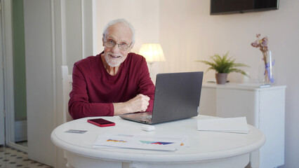 An elderly man sits at a table in front of a laptop, looks at the camera and smiles