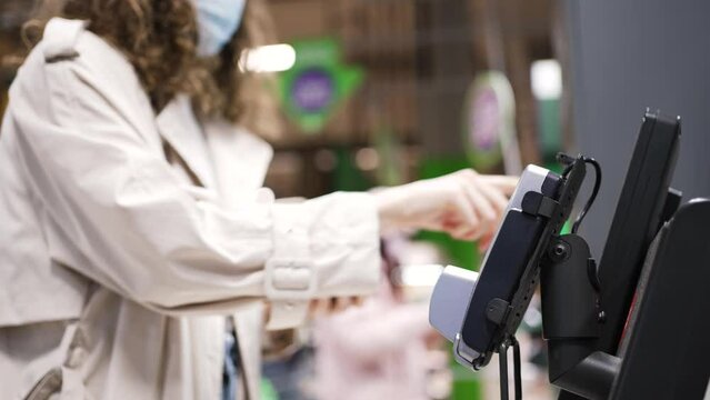 Woman in medical mask pays for products at checkout counter via app on mobile phone. Lady consumer does shopping in supermarket at quarantine closeup