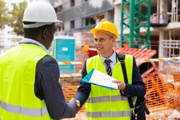 Architect and foreman, European and African-American men, standing on construction site and shaking each other hands after signing documentation.