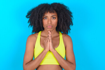 young woman with afro hairstyle in sportswear against blue background keeps palms pressed together in front of her having regretful look, asking for forgiveness. Forgive me please.