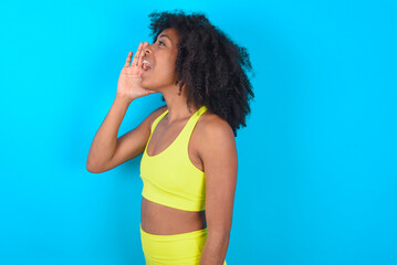 Fototapeta na wymiar young woman with afro hairstyle in sportswear against blue background profile view, looking happy and excited, shouting and calling to copy space.