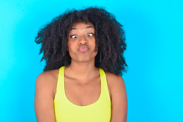 young woman with afro hairstyle in sportswear against blue background crosses eyes, puts lips, makes grimace with awkward expression has fun alone, plays fool.