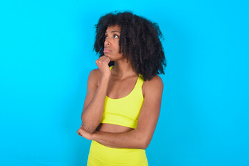 Portrait of thoughtful young woman with afro hairstyle in sportswear against blue wall keeps hand under chin, looks away trying to remember something or listens something with interest. Youth concept.