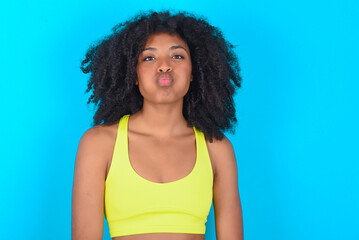 young woman with afro hairstyle in sportswear against blue background puffing cheeks with funny...