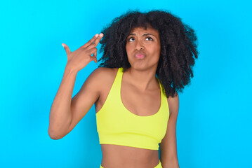 Unhappy young woman with afro hairstyle in sportswear against blue background imitates gun shoot...