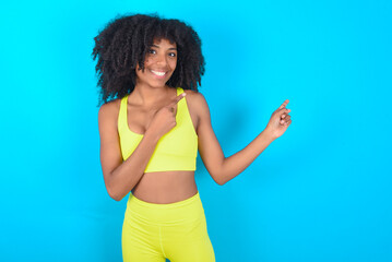 young woman with afro hairstyle in sportswear against blue background with positive expression, indicates with fore finger at blank copy space for your promotional text or advertisement.