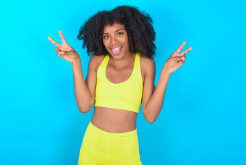 young woman with afro hairstyle in sportswear against blue background with optimistic smile, showing peace or victory gesture with both hands, looking friendly. V sign.