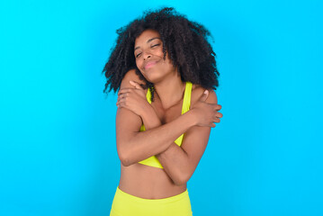 Naklejka premium young woman with afro hairstyle in sportswear against blue background Hugging oneself happy and positive, smiling confident. Self love and self care.