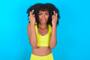 Fototapeta na wymiar young woman with afro hairstyle in sportswear against blue background holding fingers crossed with worried expression hoping boss didn't noticed mistakes at work.