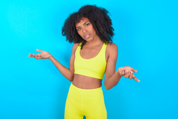 young woman with afro hairstyle in sportswear against blue background looks uncertain shrugs shoulders.