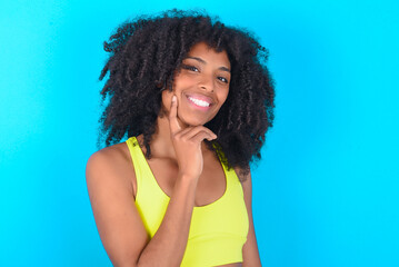 Carefree successful young woman with afro hairstyle in sportswear against blue background touching...