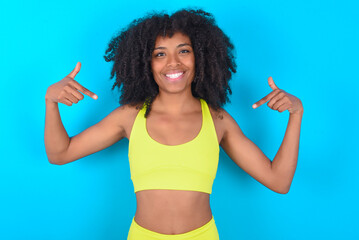 Pick me! Confident, self-assured and charismatic young woman with afro hairstyle in sportswear against blue background promoting oneself as wanting role smiling broadly and pointing at body.