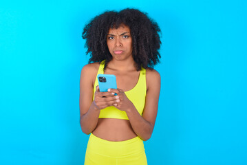 Portrait of a confused young woman with afro hairstyle in sportswear against blue background holding mobile phone and shrugging shoulders and frowning face.