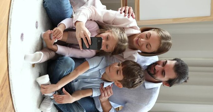 Young parents have fun use smartphone seated on floor with kids, vertical view. Mom and dad their cute children look at cellphone screen make selfie picture, enjoy new mobile app. Tech, games concept