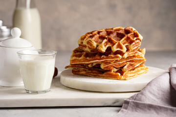 Sweet breakfast - fresh homemade belgian waffles with honey and powdered sugar stacked on white round marble board and small glass with milk