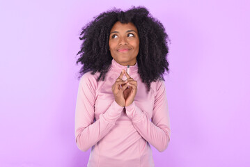 young woman with afro hairstyle in technical sports shirt against purple background steepled fingers and looks mysterious aside has great evil plan in mind