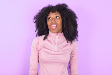 Funny young woman with afro hairstyle in technical sports shirt against purple background makes...