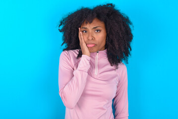 Sad lonely young woman with afro hairstyle in technical sports shirt against blue background touches cheek with hand bites lower lip and gazes with displeasure. Bad emotions