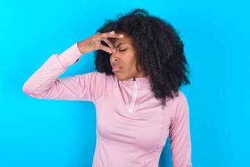 Fototapeta na wymiar Sad young woman with afro hairstyle in technical sports shirt against blue background suffering from headache holding hand on her face