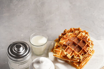 Sweet breakfast - fresh homemade belgian waffles with honey and powdered sugar stacked on white round marble board and small glass with milk, copy space