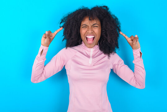 Photo of crazy young woman with afro hairstyle in technical sports shirt against blue background  screaming and pointing with fingers at hair closed eyes