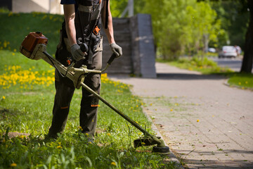 A man mows the grass in a grassy field along a footpath on a warm sunny day. A man in protective clothing, and gloves with a trimmer. - 496710371