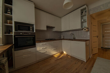 interior of a small cozy kitchen with wooden floor and white furniture, sunbeams in the room.