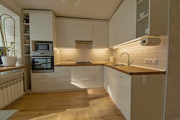 interior of a small cozy kitchen with wooden floor and white furniture, sunbeams in the room.
