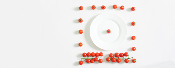 Serving red cherry tomatoes on a white kitchen table next to a round white plate. Shades of white and red. Copy space