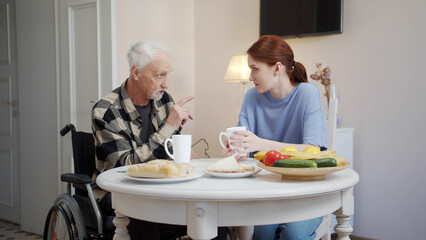 A woman communicates with a man in a wheelchair in a nursing home and drinks tea with him