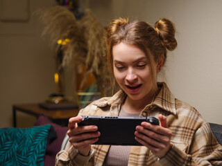 Surprised girl gamer plays video games on a portable game console in the room. Virtual reality, cyberspace, mobile games, youth culture, entertainment, recreation, game strategy.