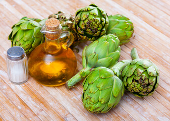 Pile of artichokes, oil bottle and saltcellar on wooden table.