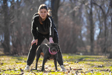 Young woman in sports costume holds active American pit bull terrier by cute pink collar, legs wide apart. Pet train or walk in park. Owner holds dog so that it does not run away without leash