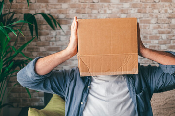 Mature man wearing empty carton box sitting on sofa in the living room of house. Caucasian guy with face hidden or covered in cardboard delivery container while relaxing on couch at home
